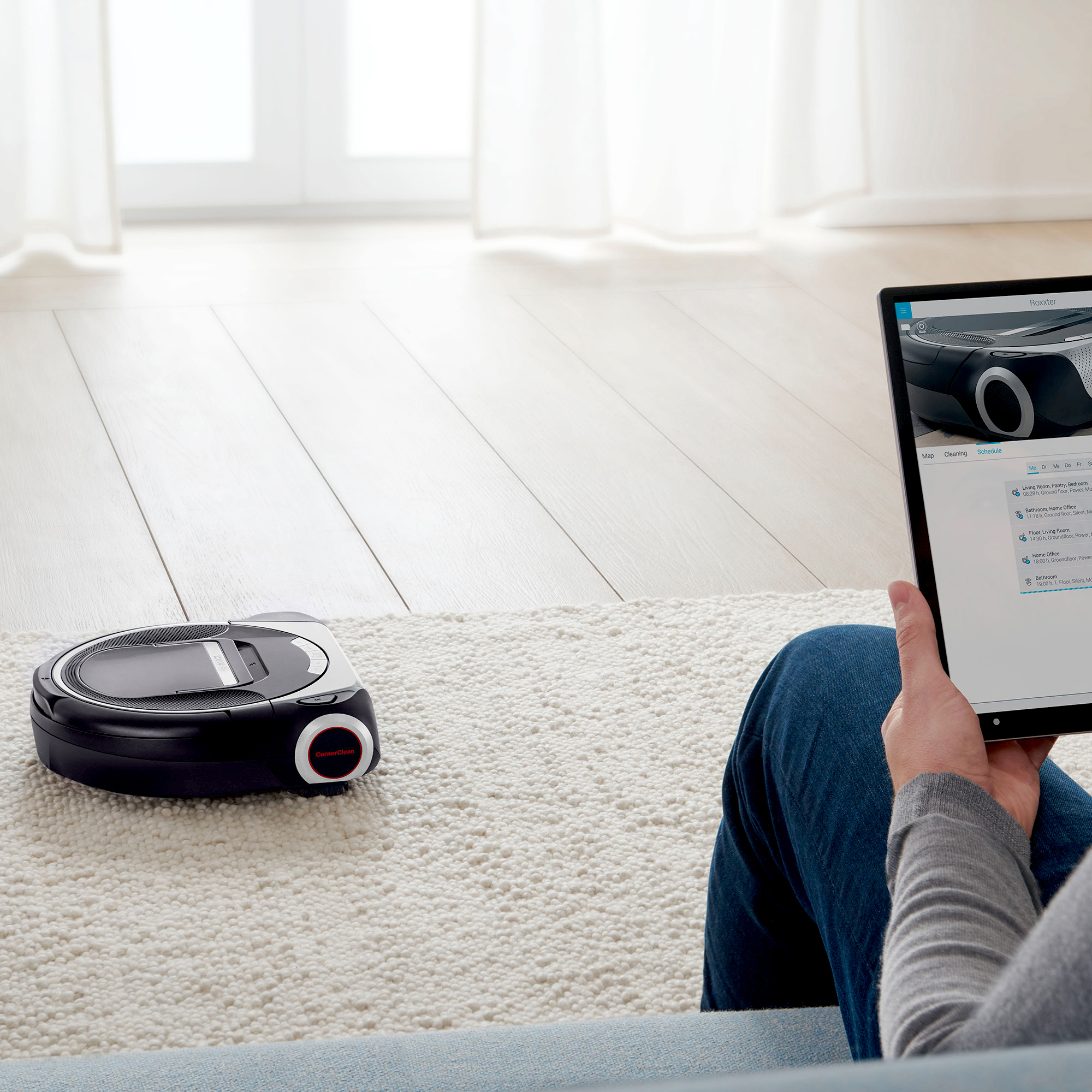 Ein schwarzer Roboter-Staubsauger auf einem weißen Teppich. Eine Person hält ein Tablet mit dem Zeitplan des Roboters.