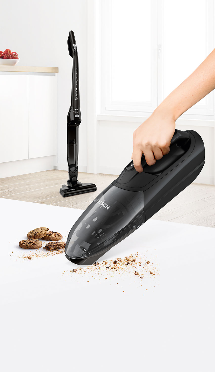 A black handheld vacuum cleaner being used to clean crumbs from a white surface. Another vacuum stands behind it.