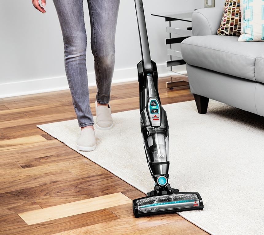 Woman vacuuming a white rug with a black and teal stick vacuum. Wooden floor.