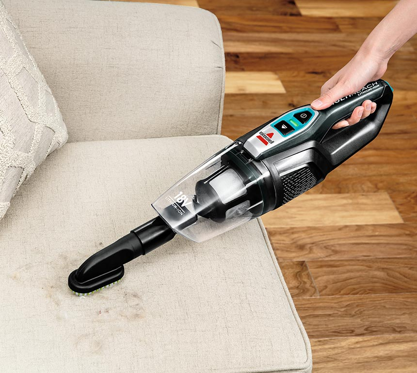 A hand holding a black and teal Bissell hand vacuum over a beige sofa with some debris on it.