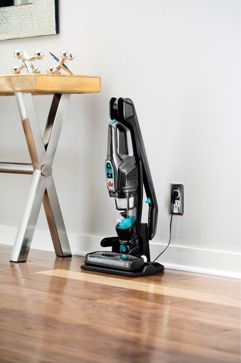 A vacuum cleaner standing upright next to a wall. A small wooden table and decor are in the background.