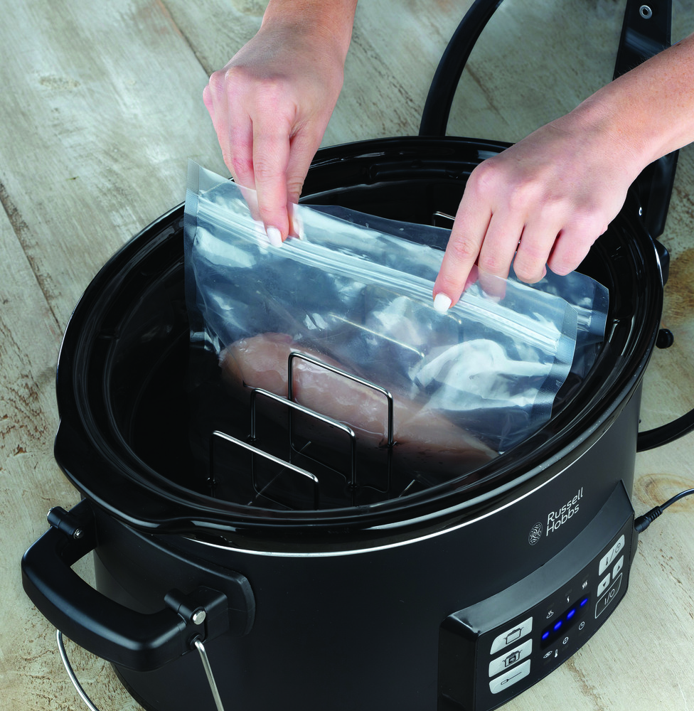 Hands putting a bag of food into a black slow cooker. Black background.
