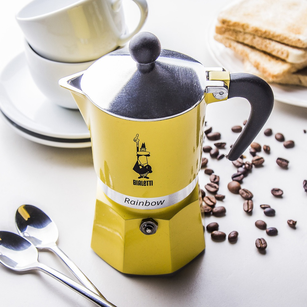 Sárga Bialetti Rainbow kávéfőző kávébabbal, kanállal és csészékkel. A kávéfőző fehér alapon van.