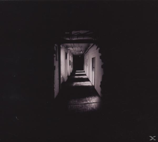 Un couloir sombre avec de la lumière au bout. Les murs et le plafond sont blancs, le sol est en bois.