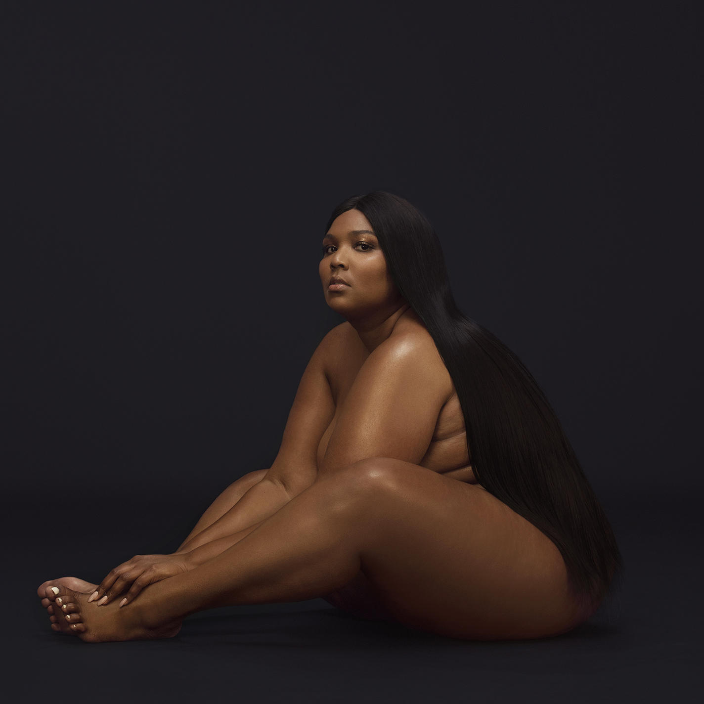 Lizzo nackt sitzend, nach oben blickend. Langes schwarzes Haar, dunkler Hintergrund. Studioaufnahme, einfach.