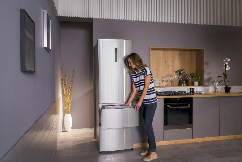 Mujer en cocina abriendo cajón del refrigerador. Refrigerador de acero inoxidable, paredes grises, detalles en madera.