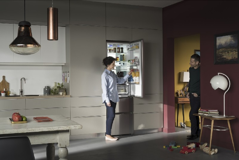 Mujer en la nevera con hombre al fondo. Ambiente de cocina, nevera abierta con comida. Pared roja de acento.