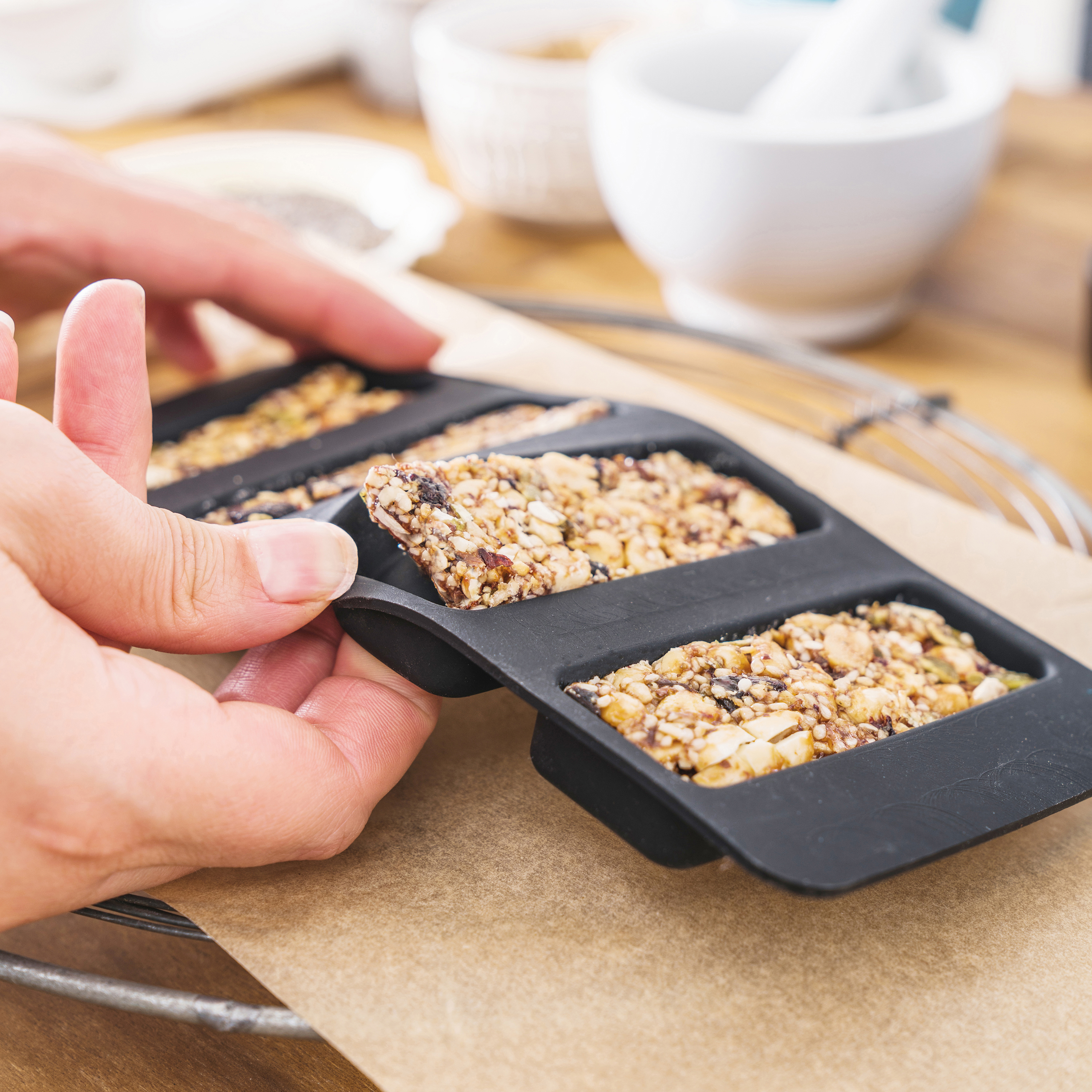 Hände halten einen Müsliriegel in einer schwarzen Silikonform, auf braunem Papier, mit anderen Zutaten.