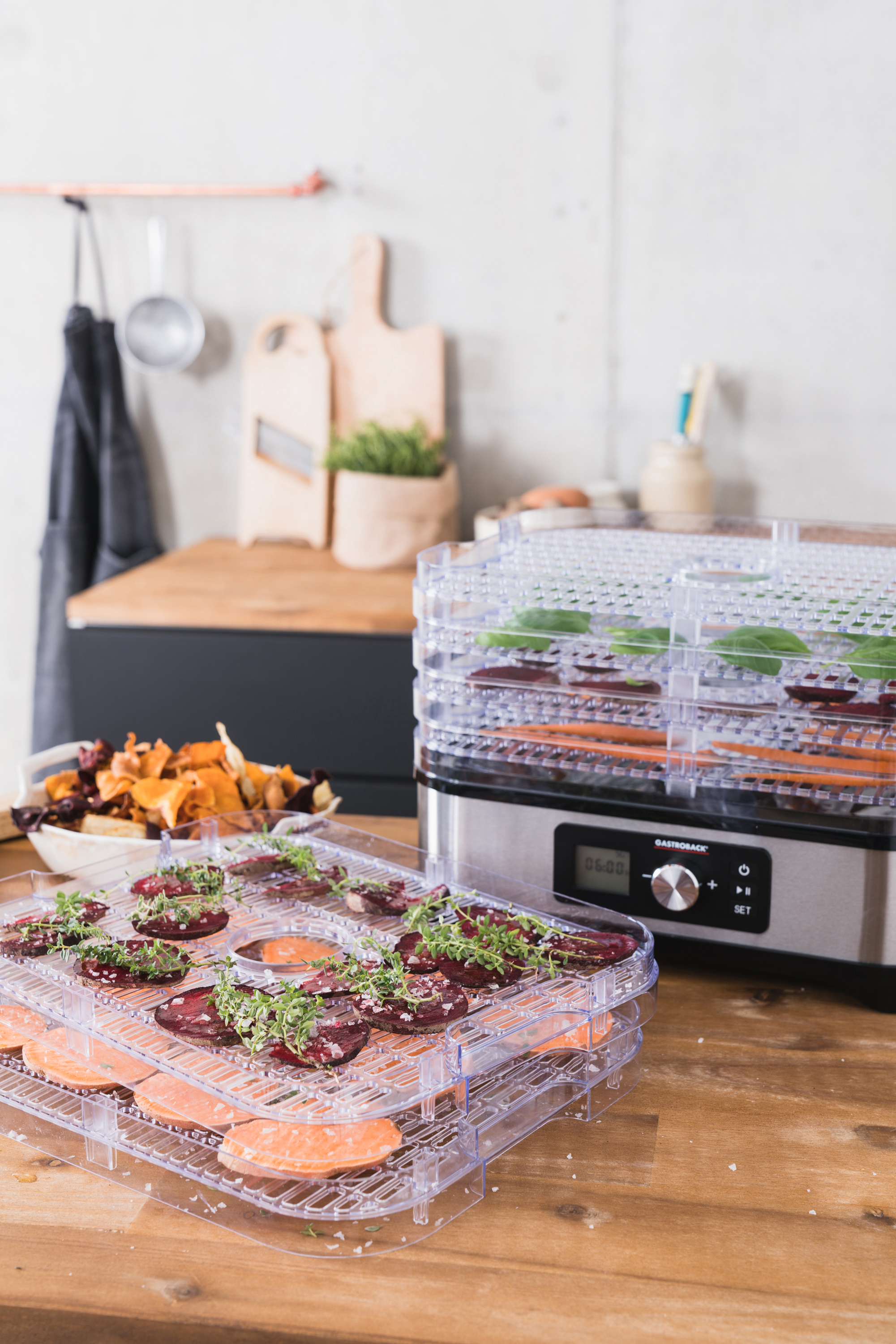 Lebensmitteltrockner mit Tabletts mit geschnittenem Gemüse und Kräutern. Ein Holztisch mit Zutaten. Heller Hintergrund.