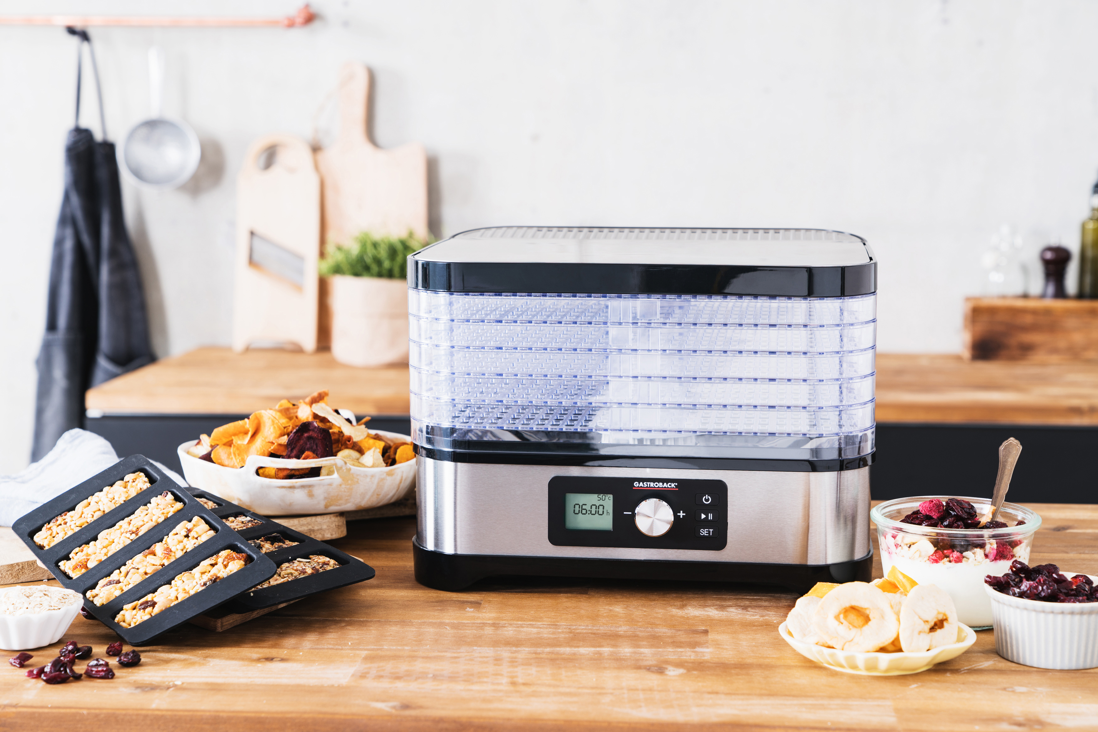 Ein Lebensmittel-Dehydrator mit Essen im Inneren und Lebensmitteln auf der Theke. Es steht auf einer Holzoberfläche.