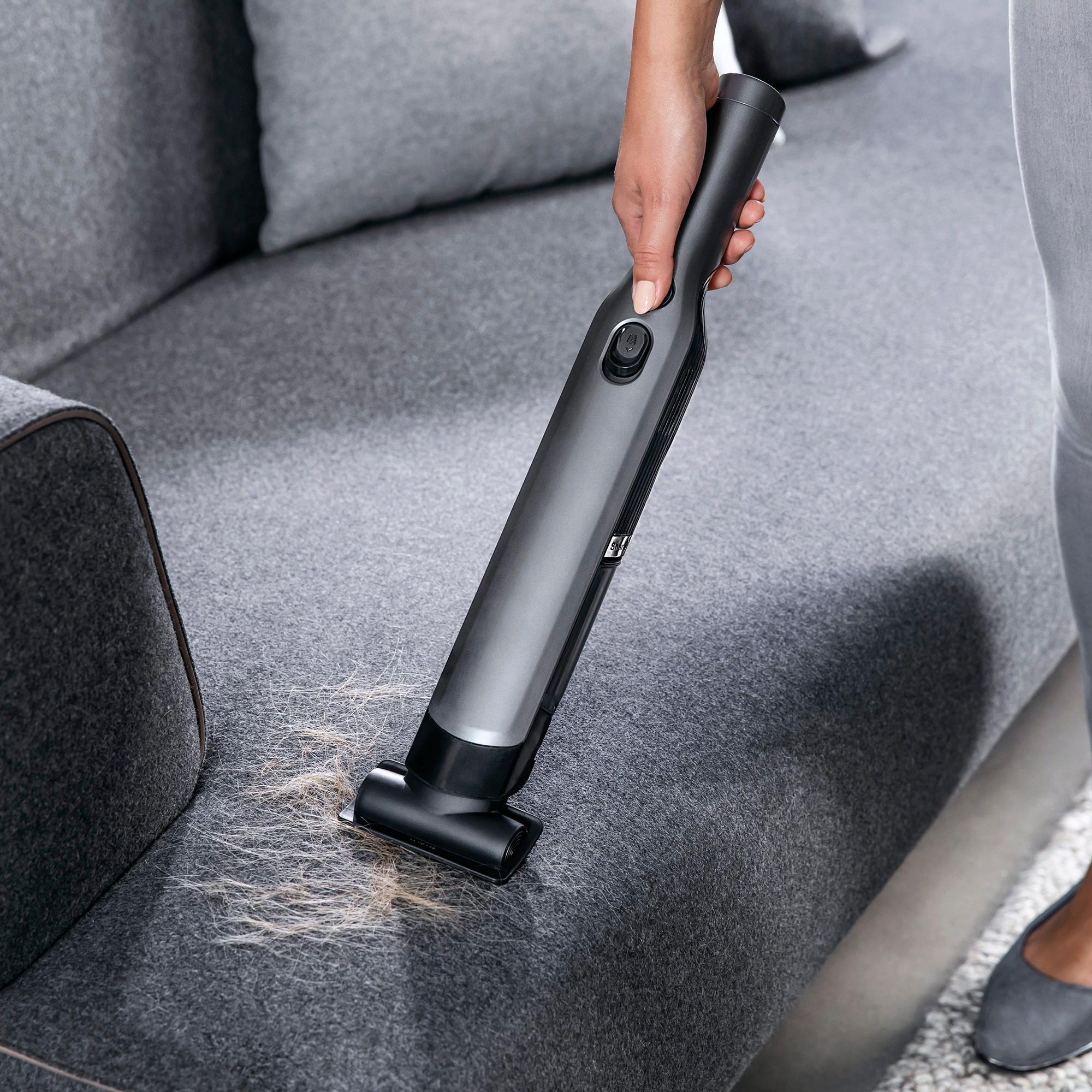 Hand saugt graues Sofa, entfernt Tierhaare. Das Sofa ist strukturiert und gut beleuchtet.