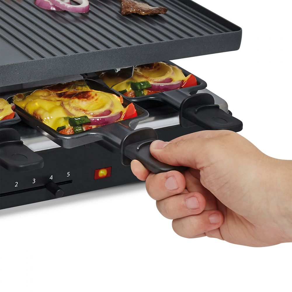 Eine Hand hält einen Raclette-Griff. Mit Essen gefüllte Pfannen im Vordergrund. Zwiebelscheiben und Fleisch auf dem Grill.