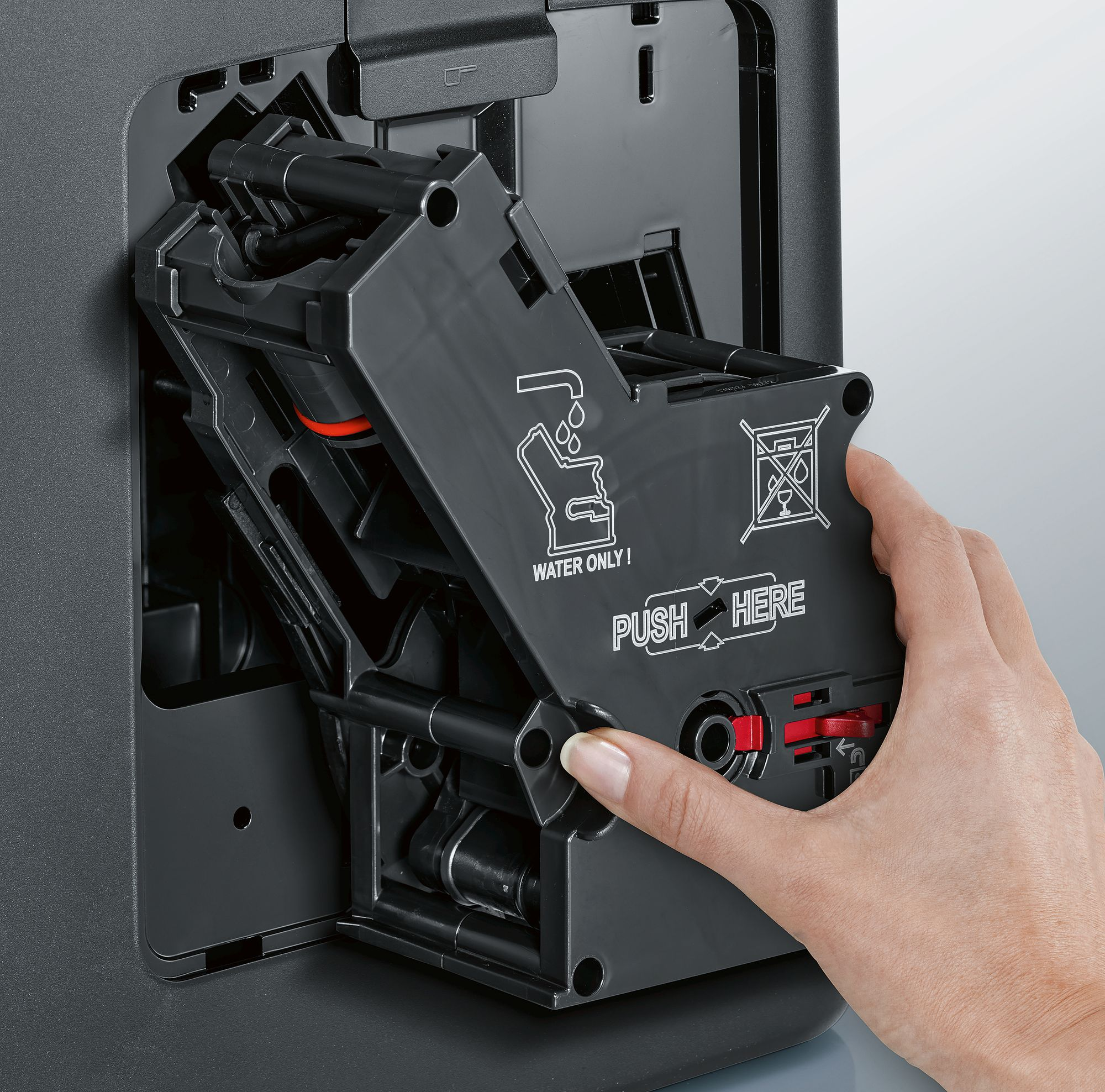 Eine Hand öffnet eine schwarze Komponente einer Kaffeemaschine mit den Worten 'PUSH HERE'.