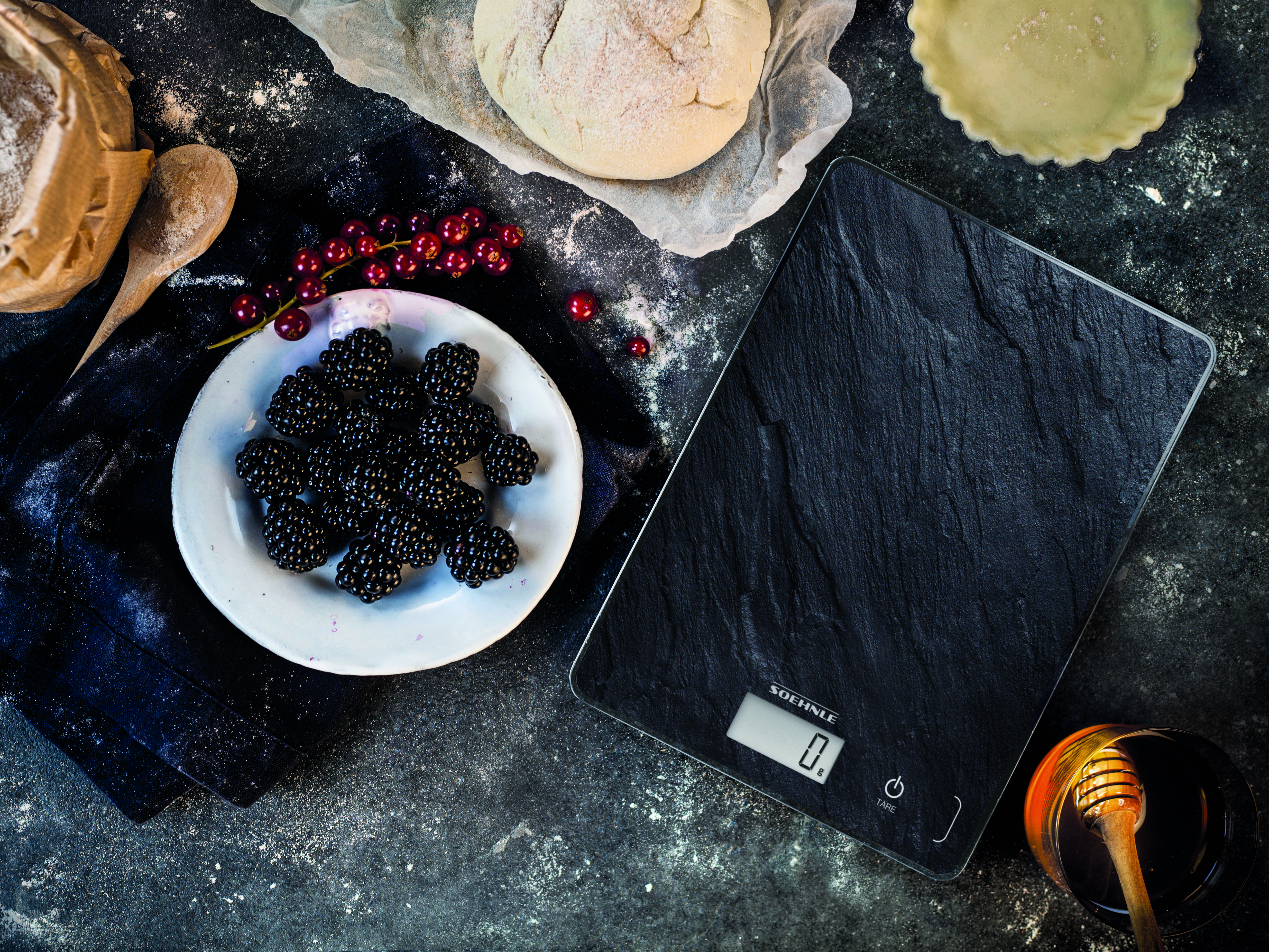 Eine schwarze digitale Küchenwaage mit Brombeeren, Honig und Zutaten auf dunkler Oberfläche.