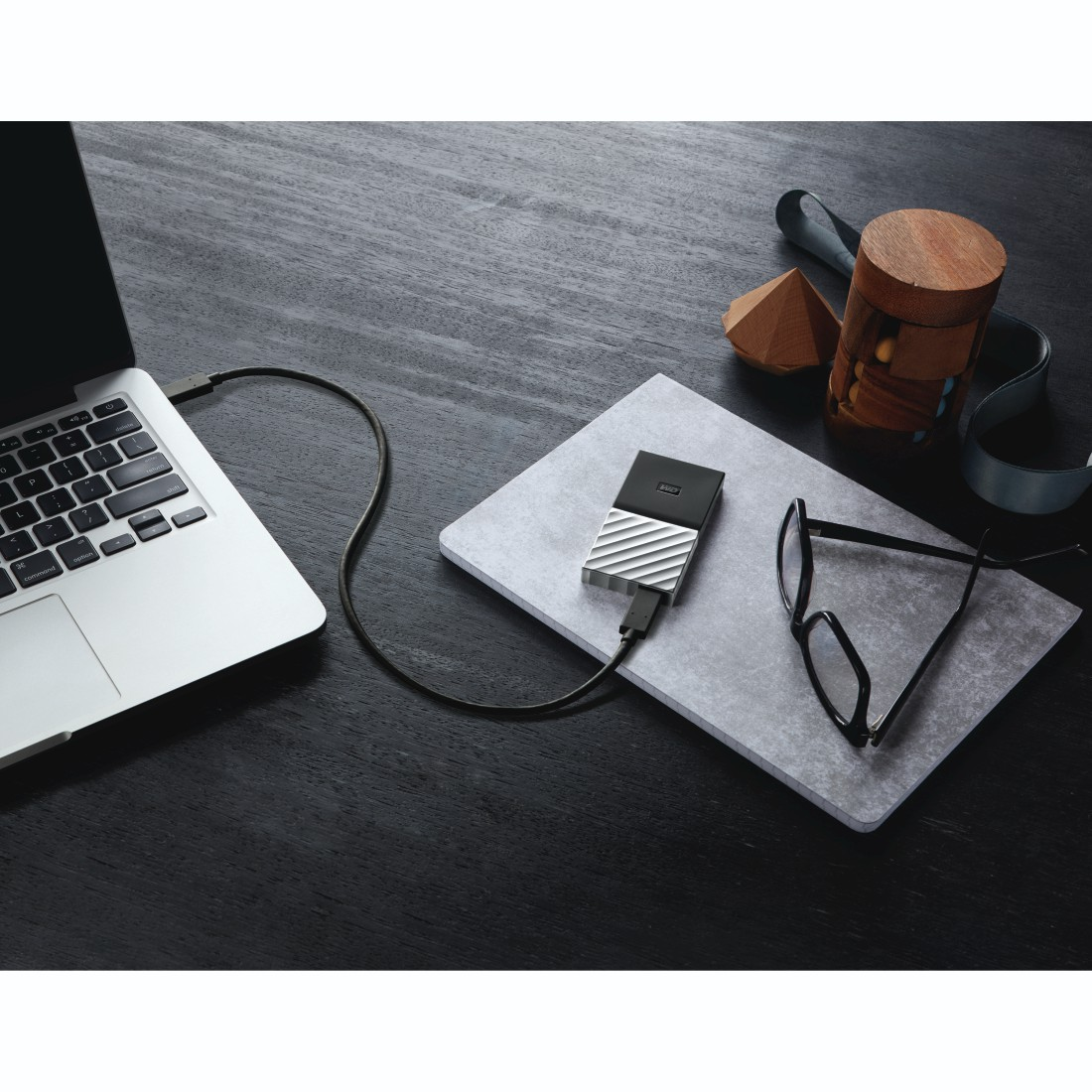 Laptop with connected external hard drive. Glasses and a notebook are placed on a light gray surface.