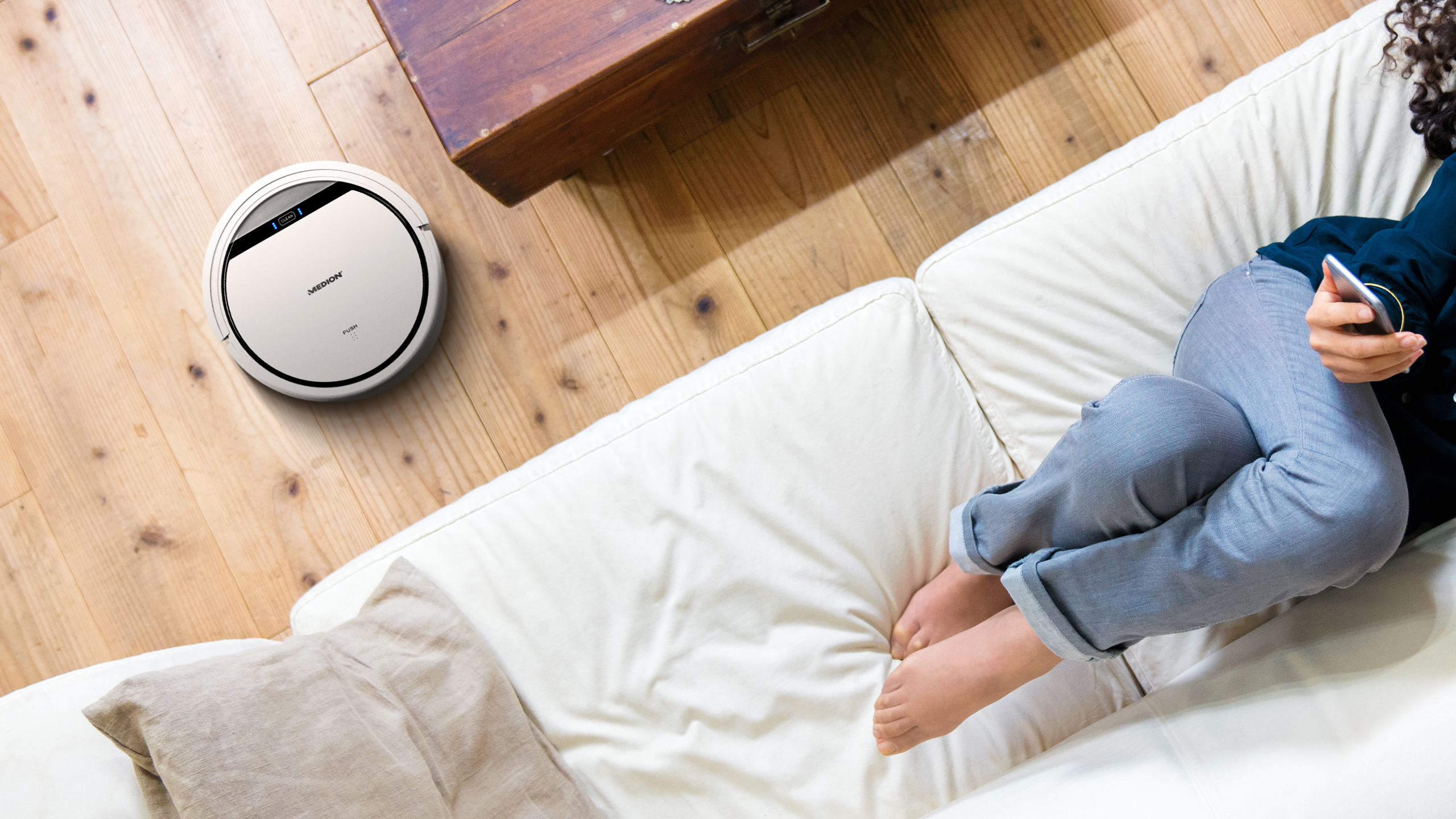 Ein weißer Roboter-Staubsauger auf einem Holzboden, neben einem weißen Sofa.