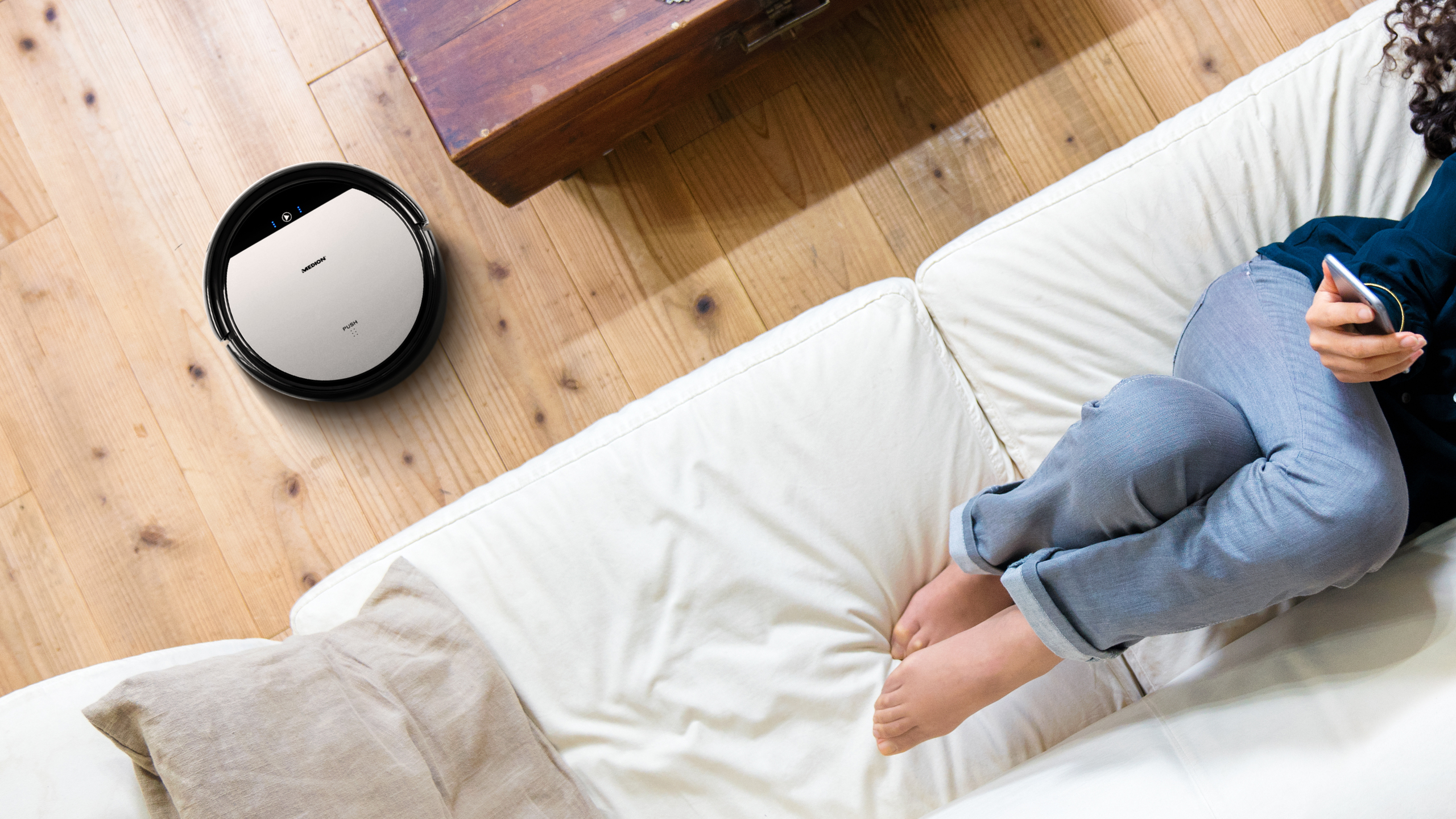 Ein Roboter-Staubsauger auf einem Holzboden. Eine Frau sitzt in der Nähe auf einem weißen Sofa.
