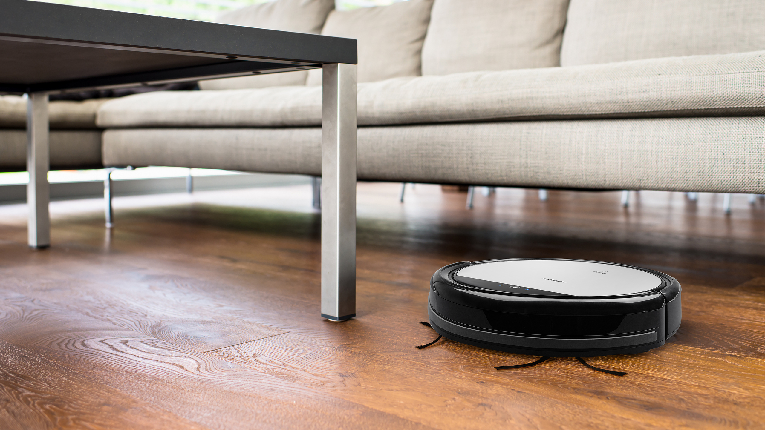 Ein schwarz-silberner Roboter-Staubsauger auf einem Holzboden in der Nähe eines Sofas und eines Tisches.