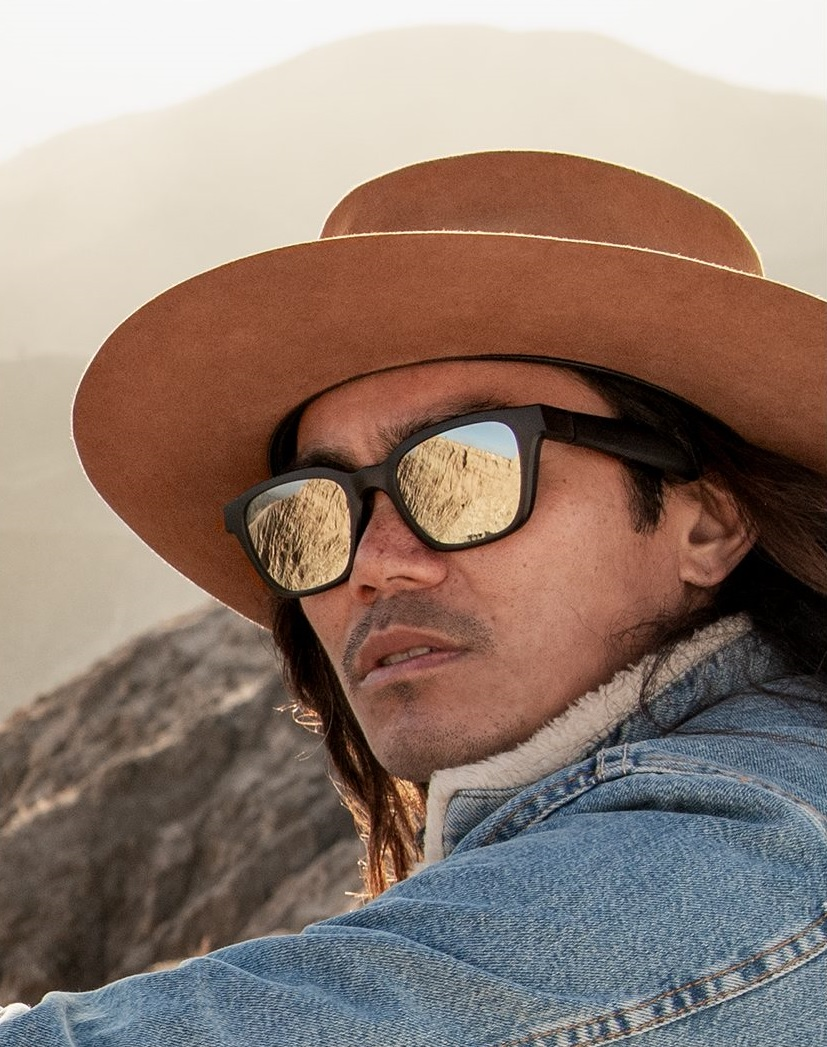 Man wearing a hat and sunglasses. The sunglasses reflect a mountain landscape.