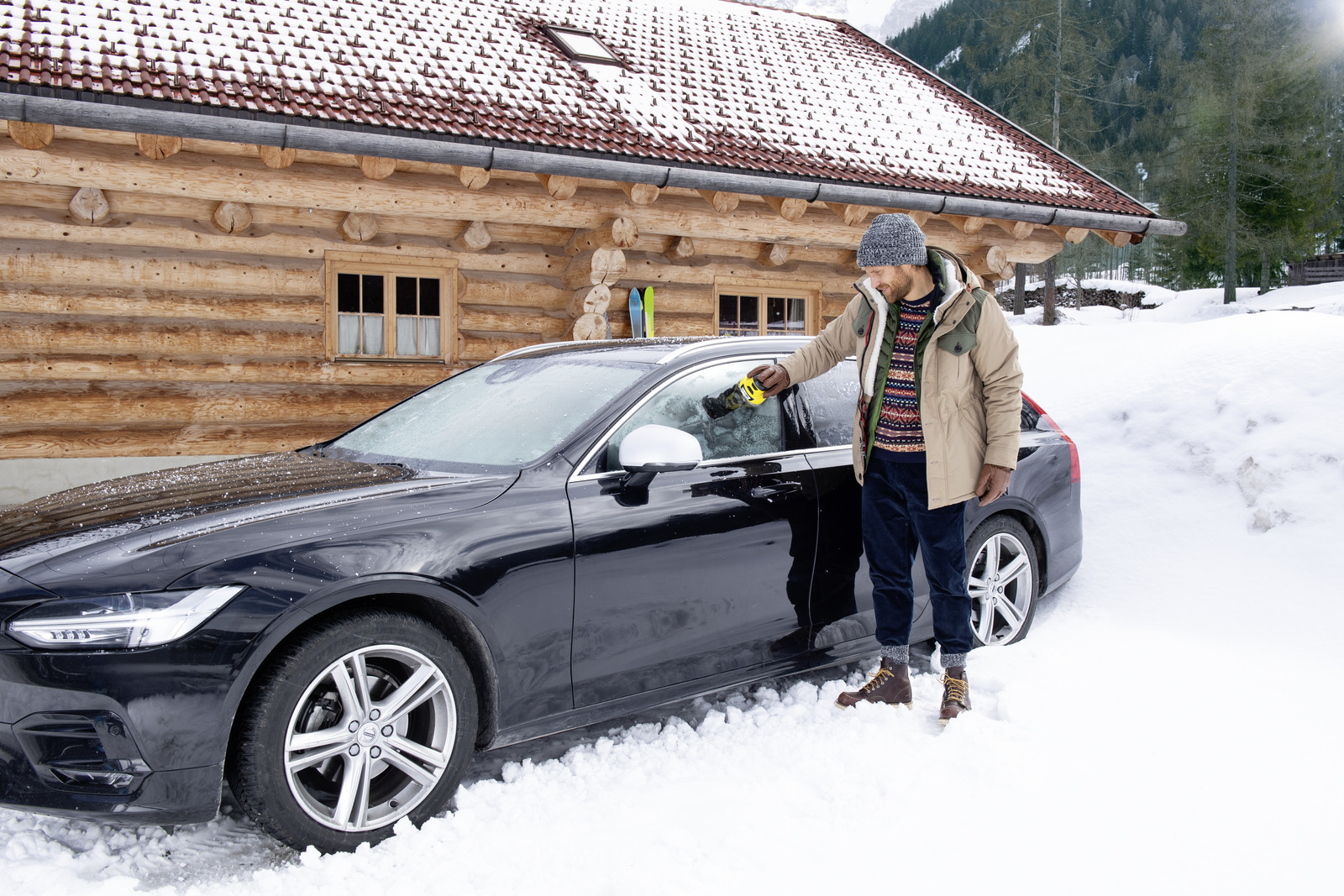 Un uomo che toglie la neve dal finestrino di un'auto in inverno. Una baita sullo sfondo.