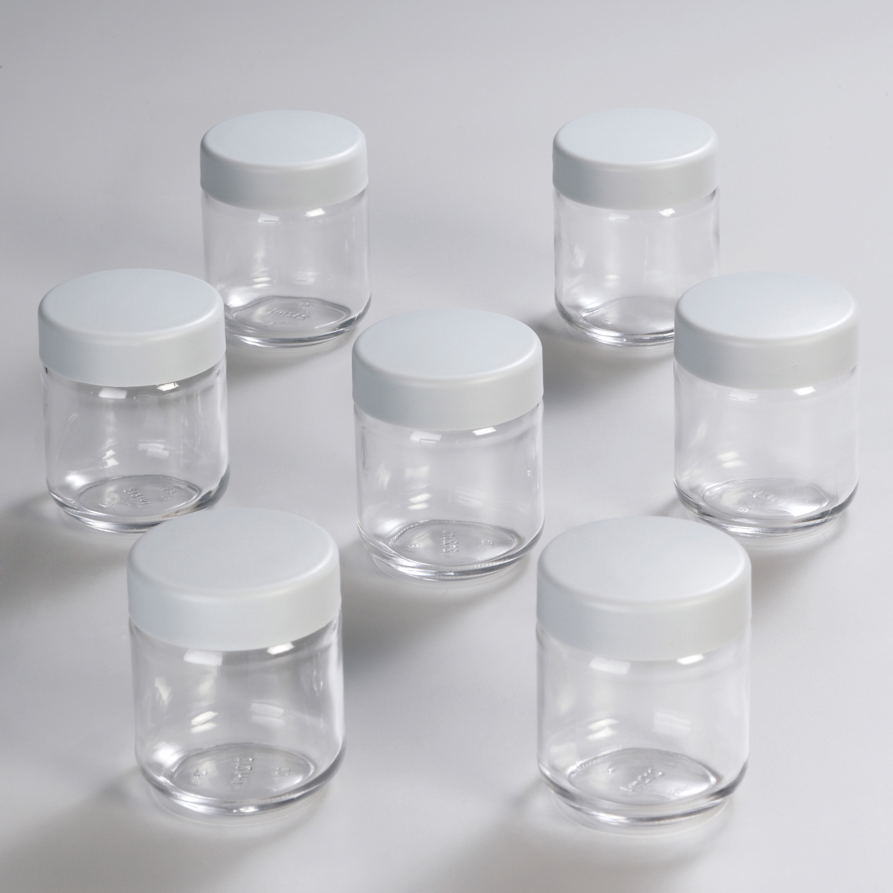 Sieben transparente Glasbehälter mit weißen Deckeln. Auf weißer Oberfläche.