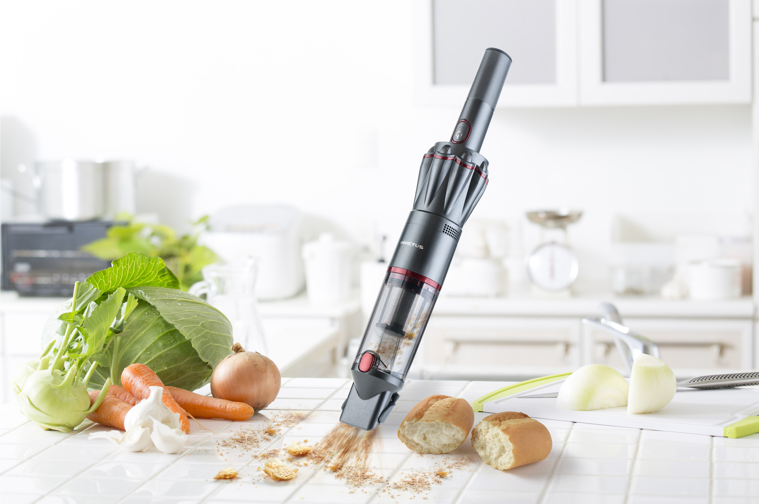 Ein Staubsauger reinigt Krümel auf einer weißen Küchenoberfläche. Gemüse, Brot und Küchenutensilien sind ebenfalls vorhanden.
