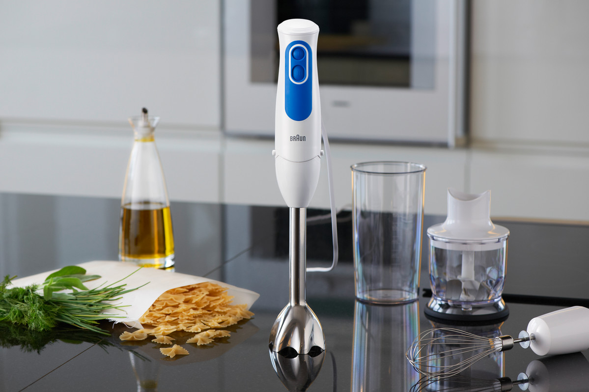 A white and blue hand blender with accessories, including a jar and whisk, with food items.