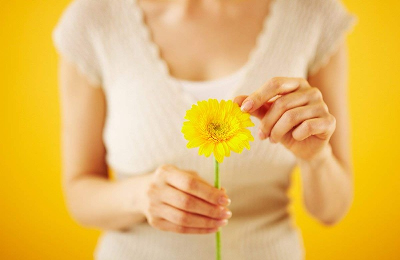 Una mujer sostiene una flor amarilla. El fondo es de color amarillo sólido.