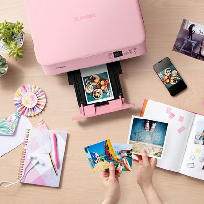 Una impresora Canon Pixma rosa imprimiendo fotos. Fotos, un teléfono y artículos de papelería en la mesa de madera.