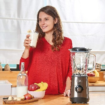 Una mujer con un suéter rojo bebiendo de un vaso, con una licuadora y fruta.