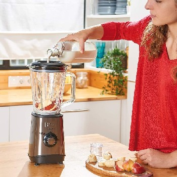 Una mujer vierte líquido con fruta en una licuadora. Fondo de cocina con armarios y encimera de madera.