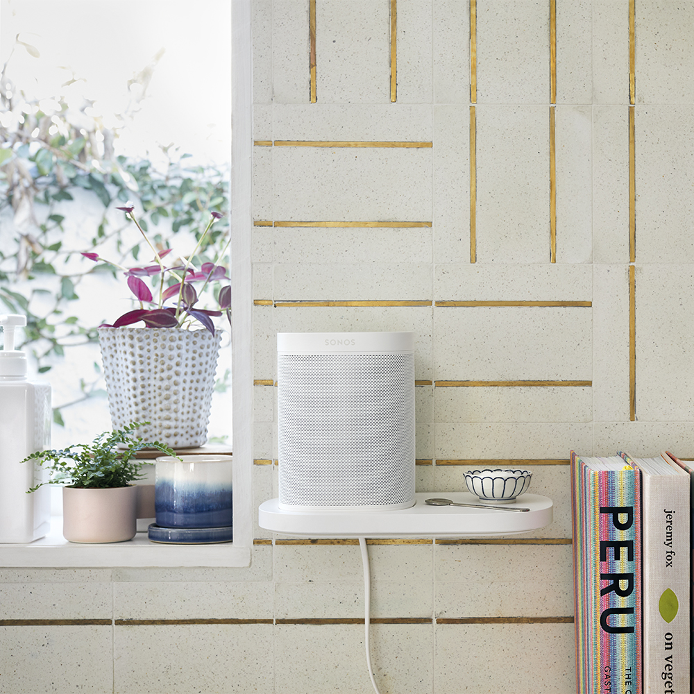 Ein weißer Sonos-Lautsprecher auf einem Regal, mit Pflanzen, Büchern und einer gefliesten Wand.