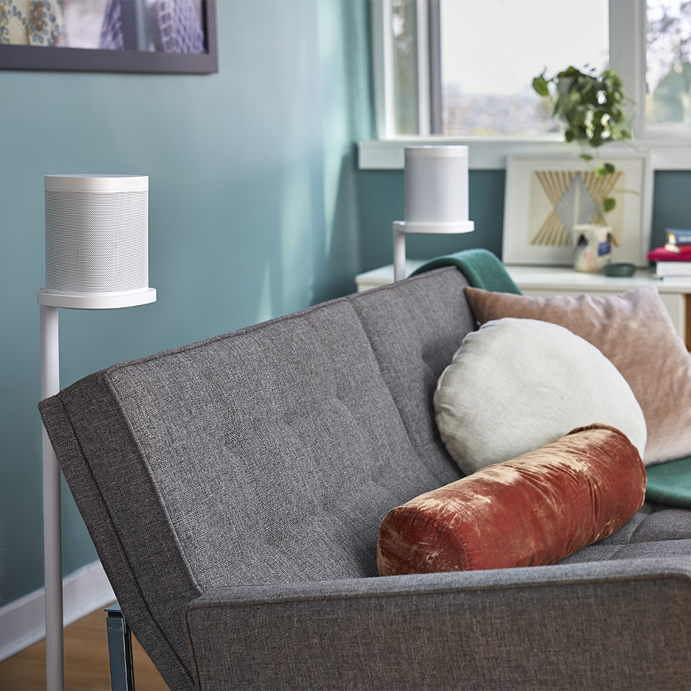 Zwei weiße Lautsprecher auf Ständern neben einem grauen Sofa mit Kissen, vor einer türkisfarbenen Wand.
