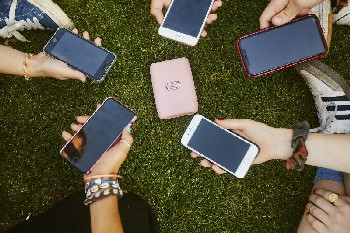 Manos sosteniendo teléfonos inteligentes en círculo sobre hierba. Un cargador inalámbrico rosa está en el centro.