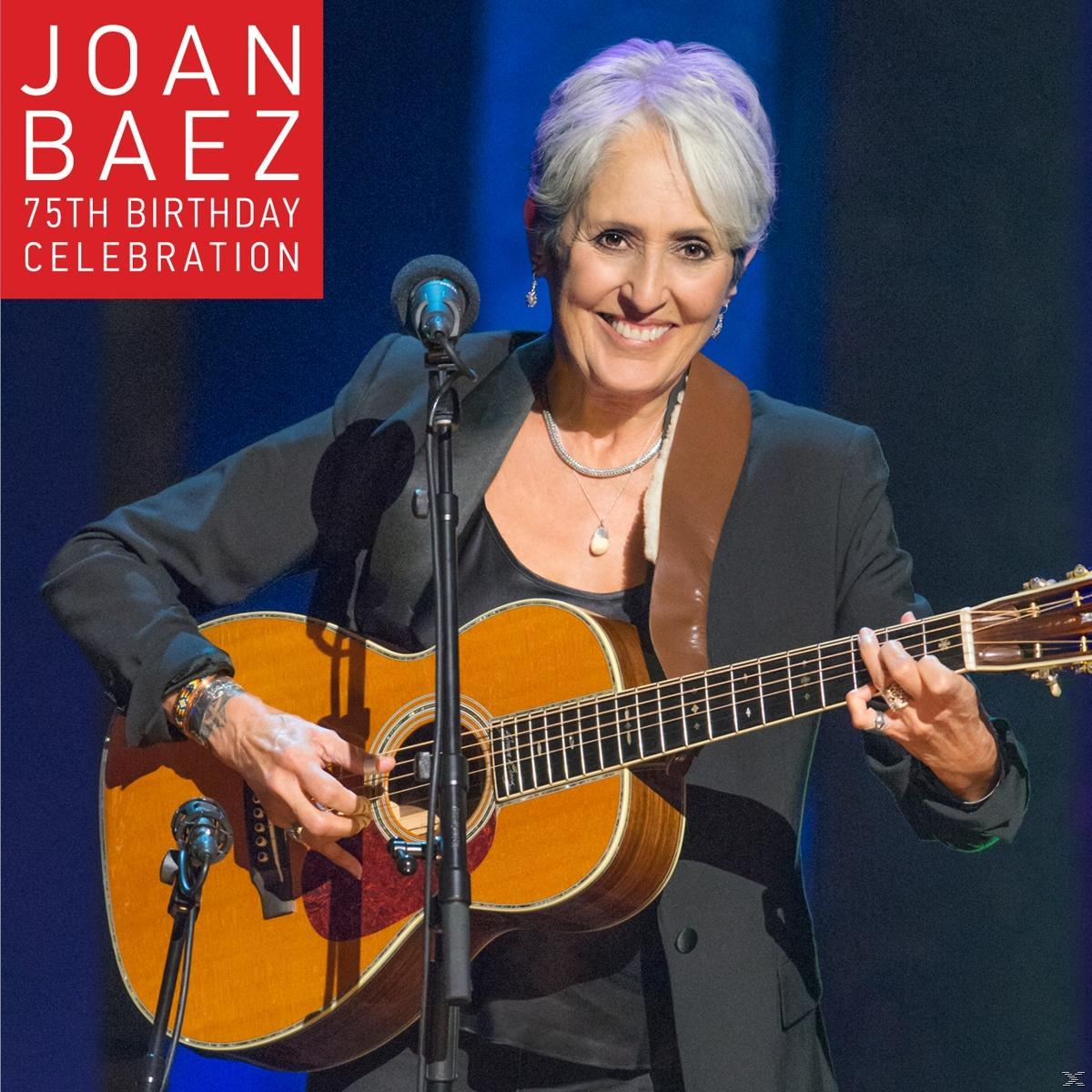 Joan Baez gitározik a színpadon, mosolyog. Fekete blézert, ékszereket visel, és egy barna gitárt tart. A színpad kék.
