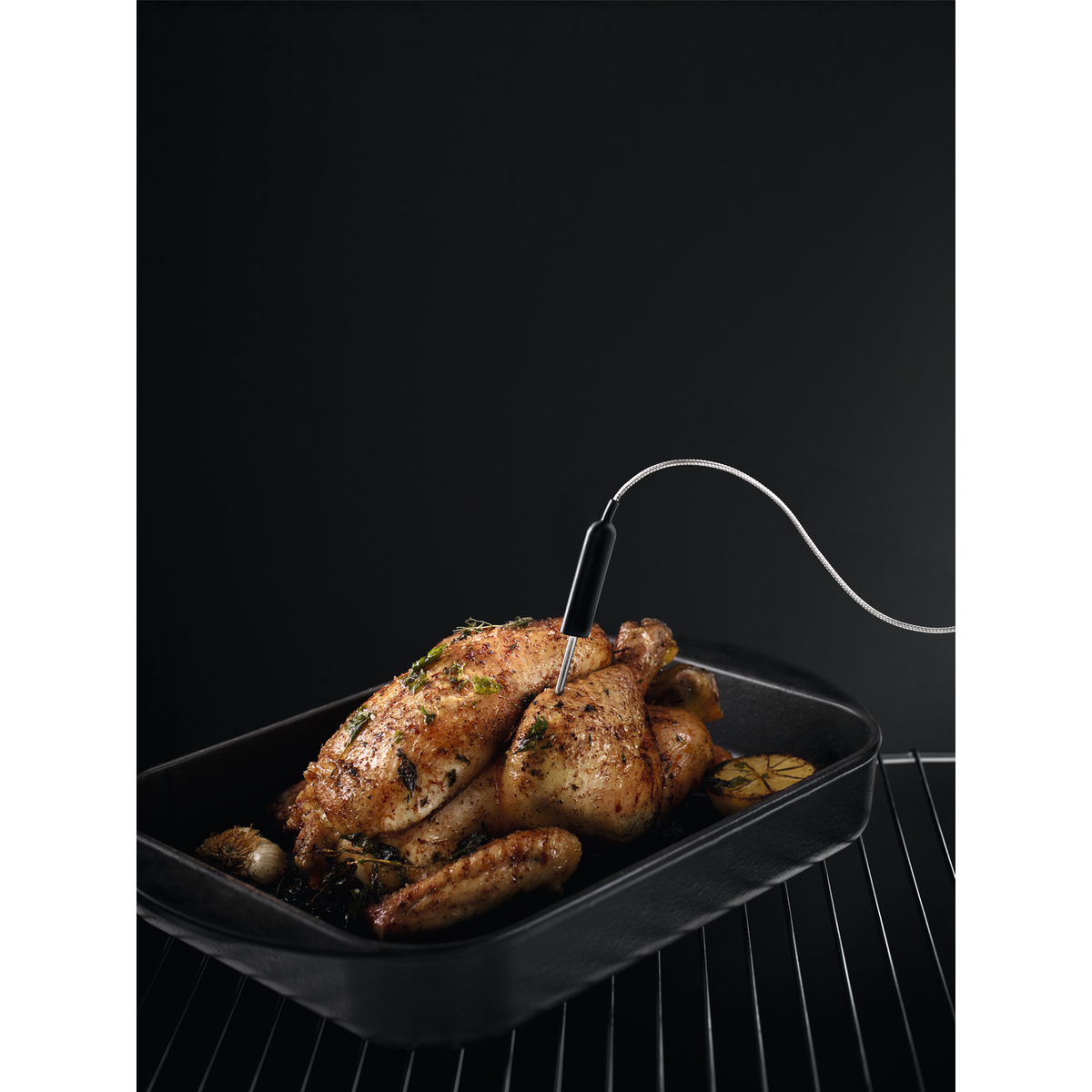 Pollo asado en una sartén negra, con un termómetro insertado. Fondo oscuro.