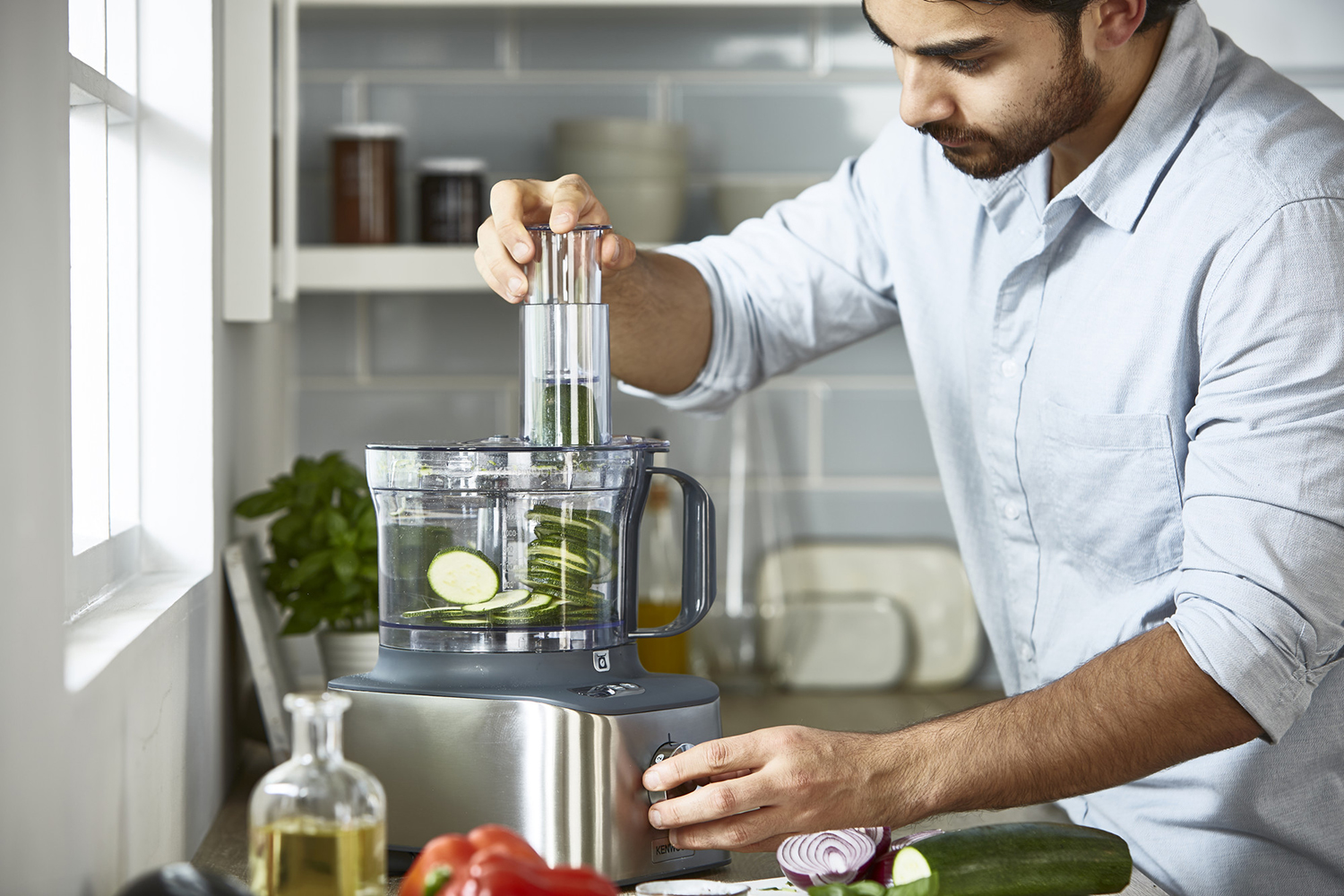 A man slicing zucchini with a food processor. Vegetables on the counter and a window in the background.