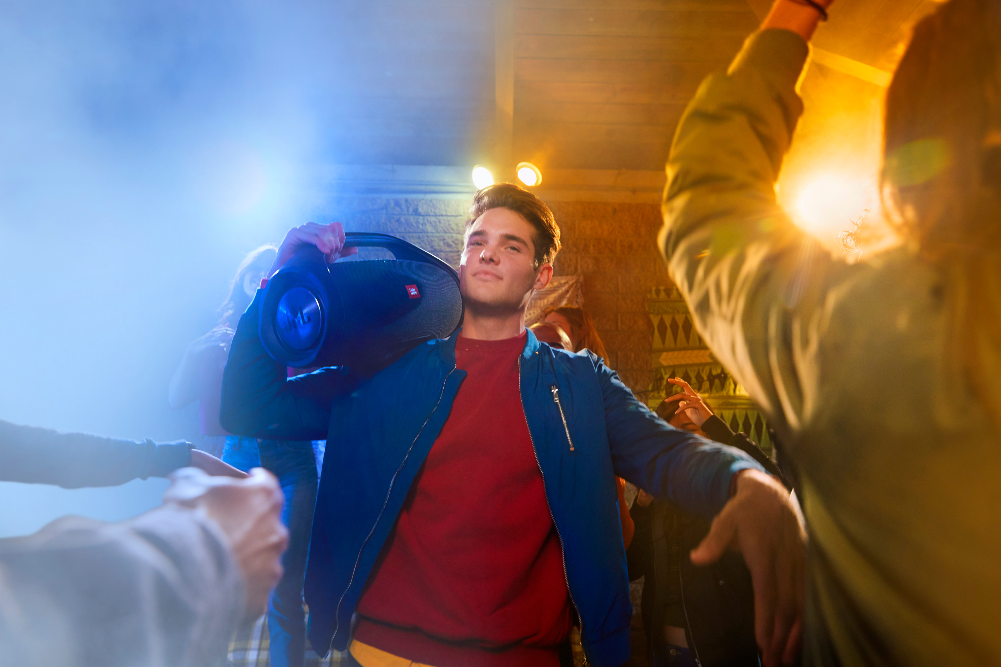 Junger Mann hält eine schwarze JBL-Boombox, lächelt auf einer Party mit tanzenden Menschen.