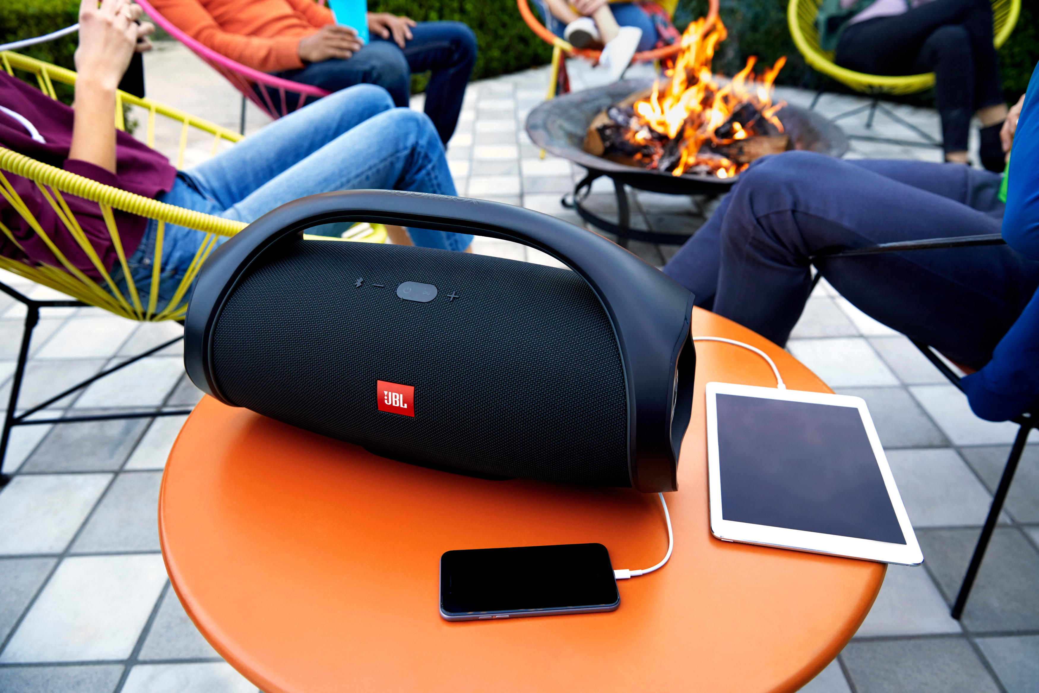 Schwarzer Lautsprecher auf einem orangen Tisch mit Telefon und Tablet. Personen und Feuer im Hintergrund.
