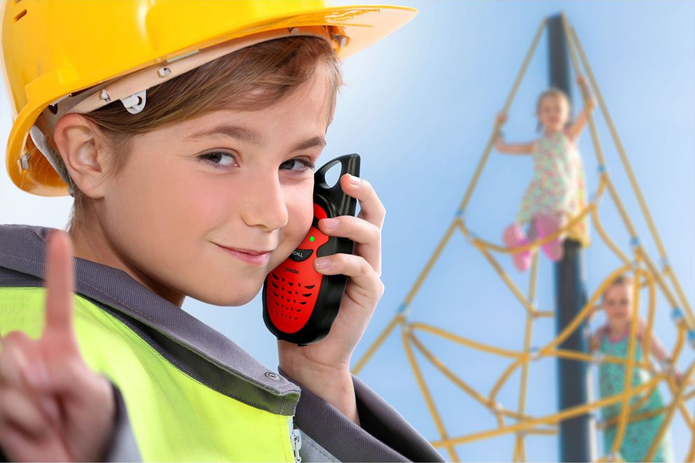 Ein Kind mit gelbem Helm hält ein Walkie-Talkie, andere Kinder spielen.