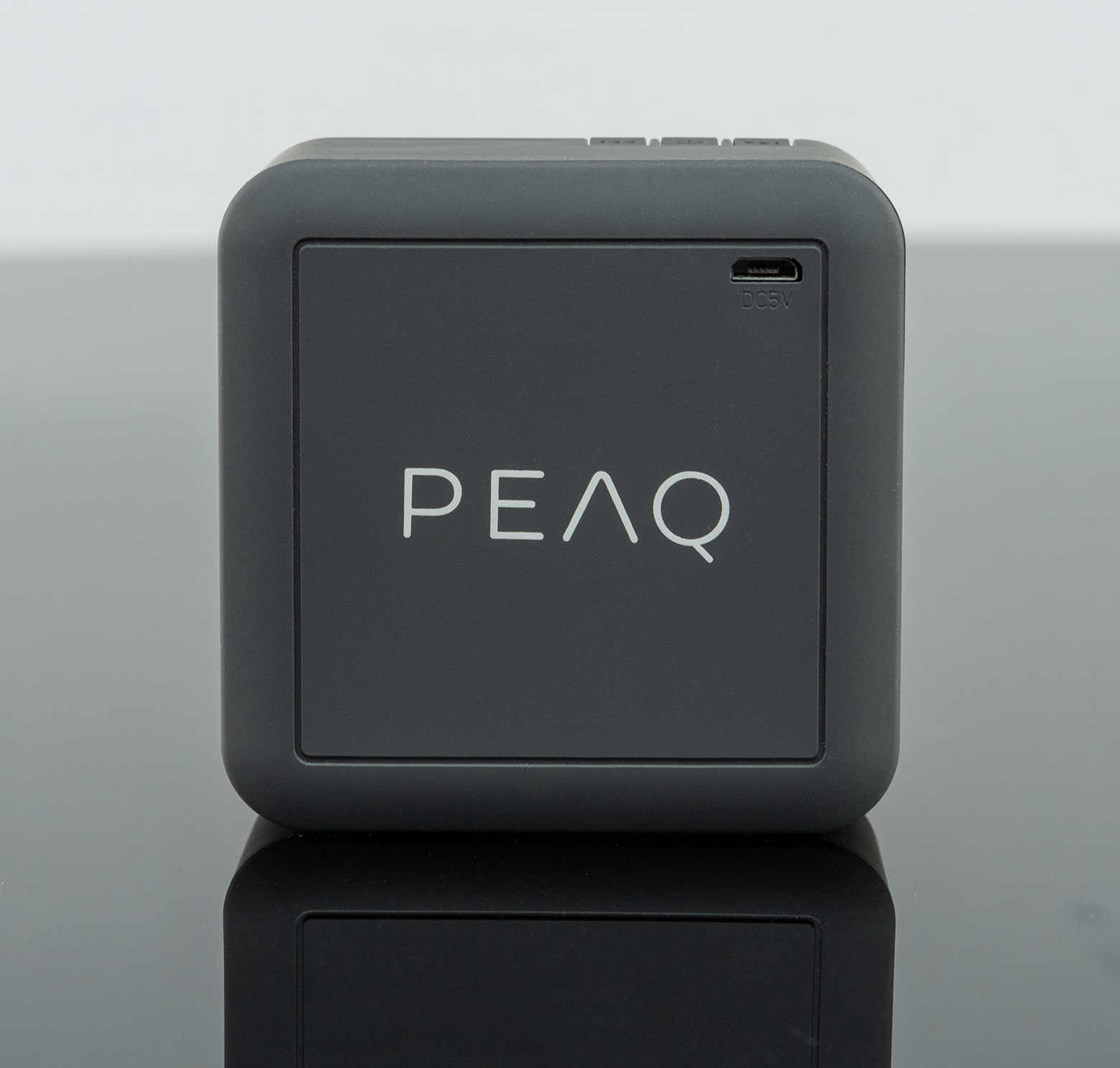 Haut-parleur gris carré avec logo PEAQ, surface noire miroitante en dessous.