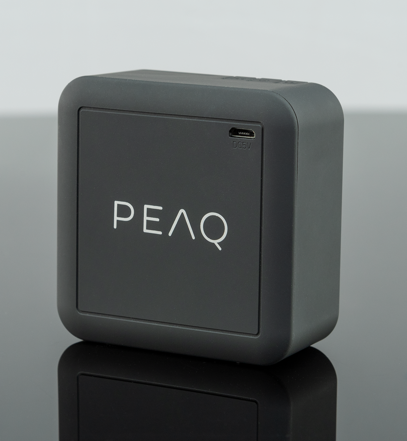 Un appareil gris foncé en forme de cube avec le mot 'PEAQ' sur le devant, sur une surface réfléchissante.