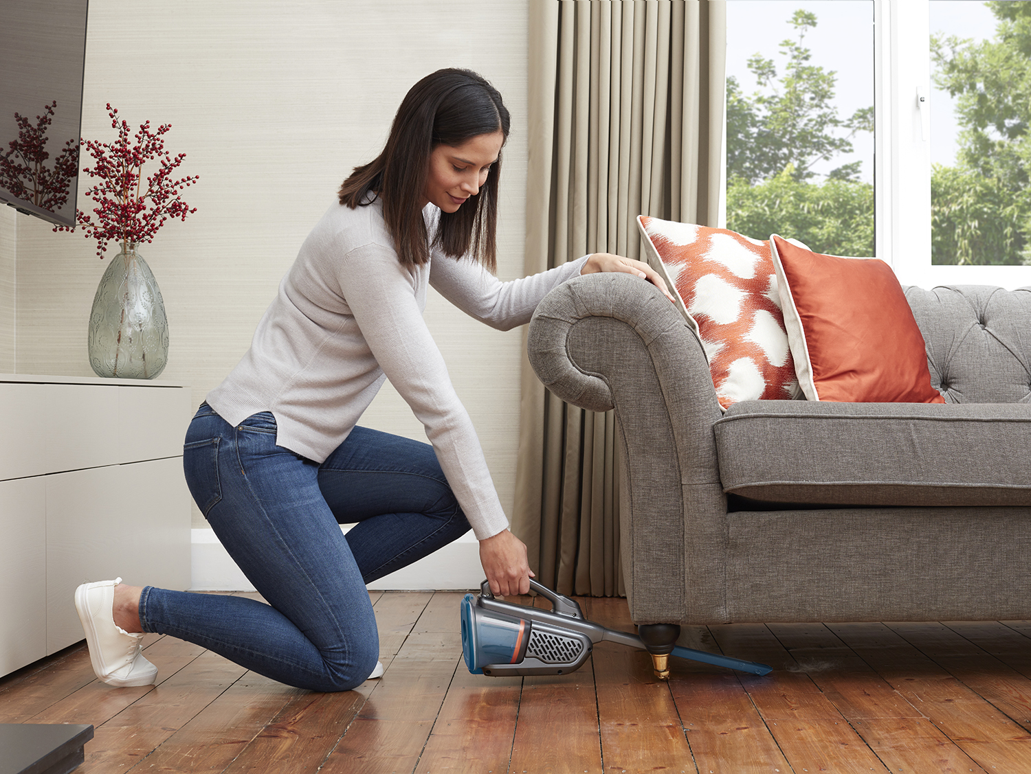 Woman cleaning near couch. She's using a handheld vacuum, with a red sofa and window in background.