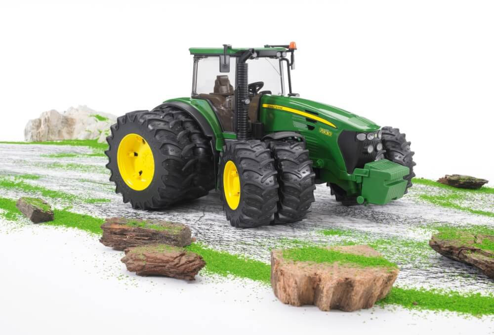 Ein grünes John Deere Traktormodell mit gelben Rädern, auf einer weißen Oberfläche mit Miniaturlandschaft aus Felsen und Gras.