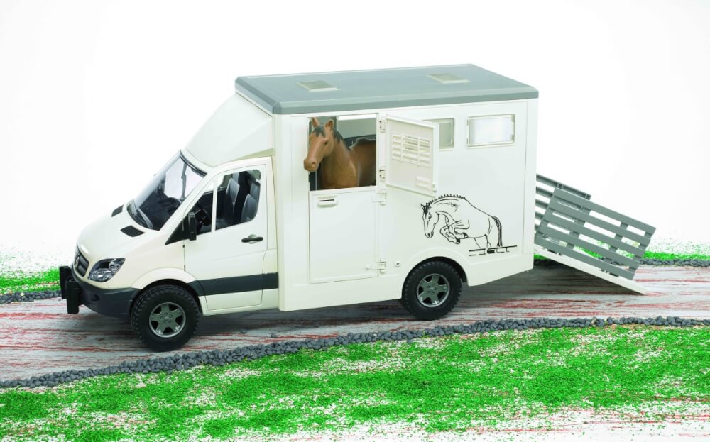 Ein weißer Pferdeanhänger mit einem Pferd darin. Der Anhänger steht auf einer Straße mit grünem Gras und kleinen Felsen an den Seiten.