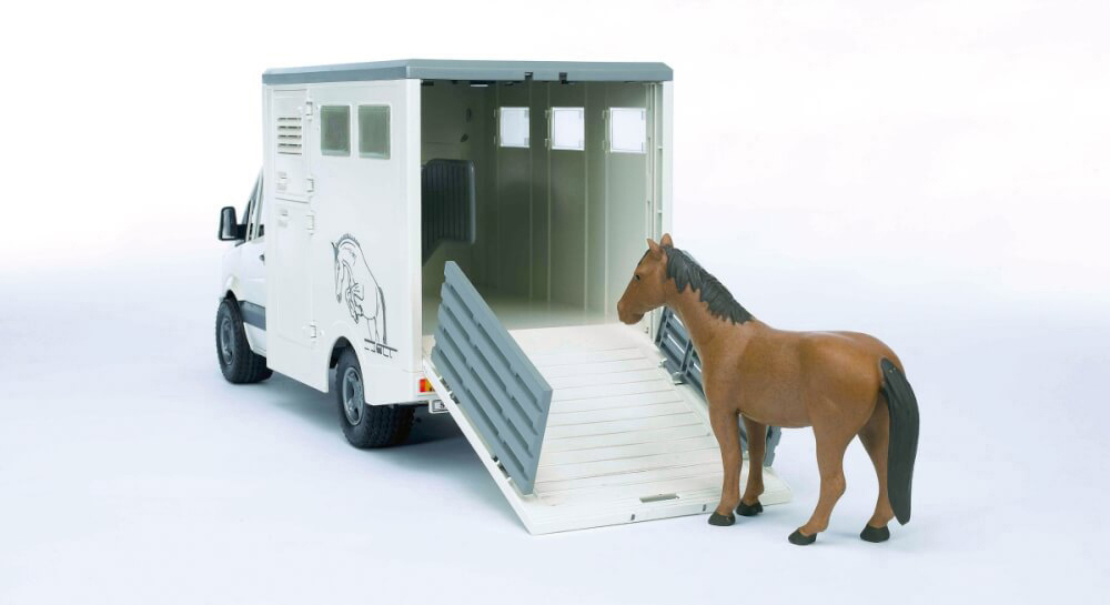 Ein Spielzeugpferd steht neben einem weißen Pferdetransporter mit offener Rampe. Das Pferd ist braun, und der Anhänger hat eine Pferdeillustration.
