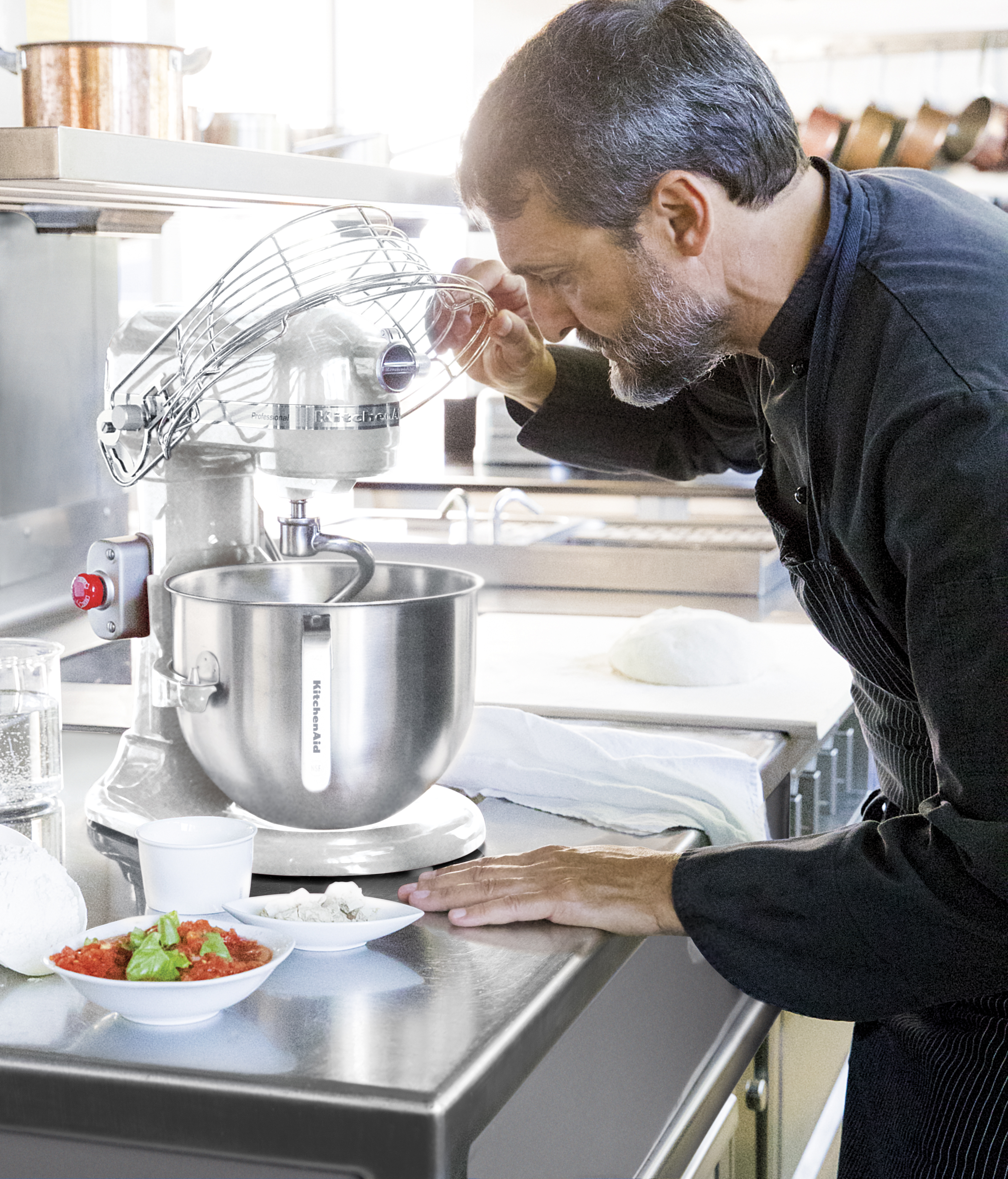 Ein Koch in schwarzer Kleidung inspiziert einen KitchenAid-Mixer. Schüsseln mit Zutaten stehen auf einer Edelstahltheke. Hintergrund: Küche.