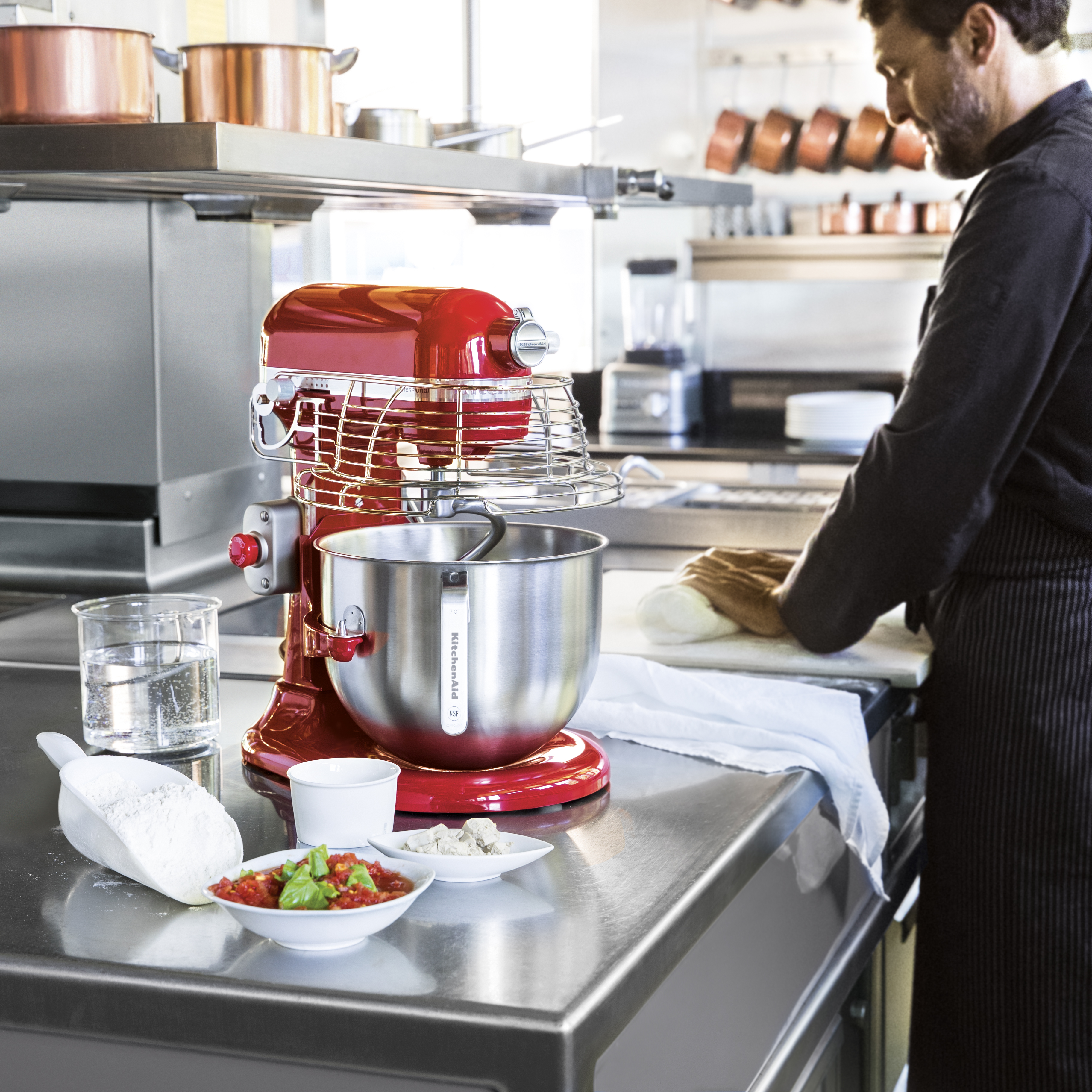 Ein roter KitchenAid-Mixer steht auf einer Theke mit Zutaten. Im Hintergrund arbeitet ein Koch an Teig. Töpfe hängen darüber.