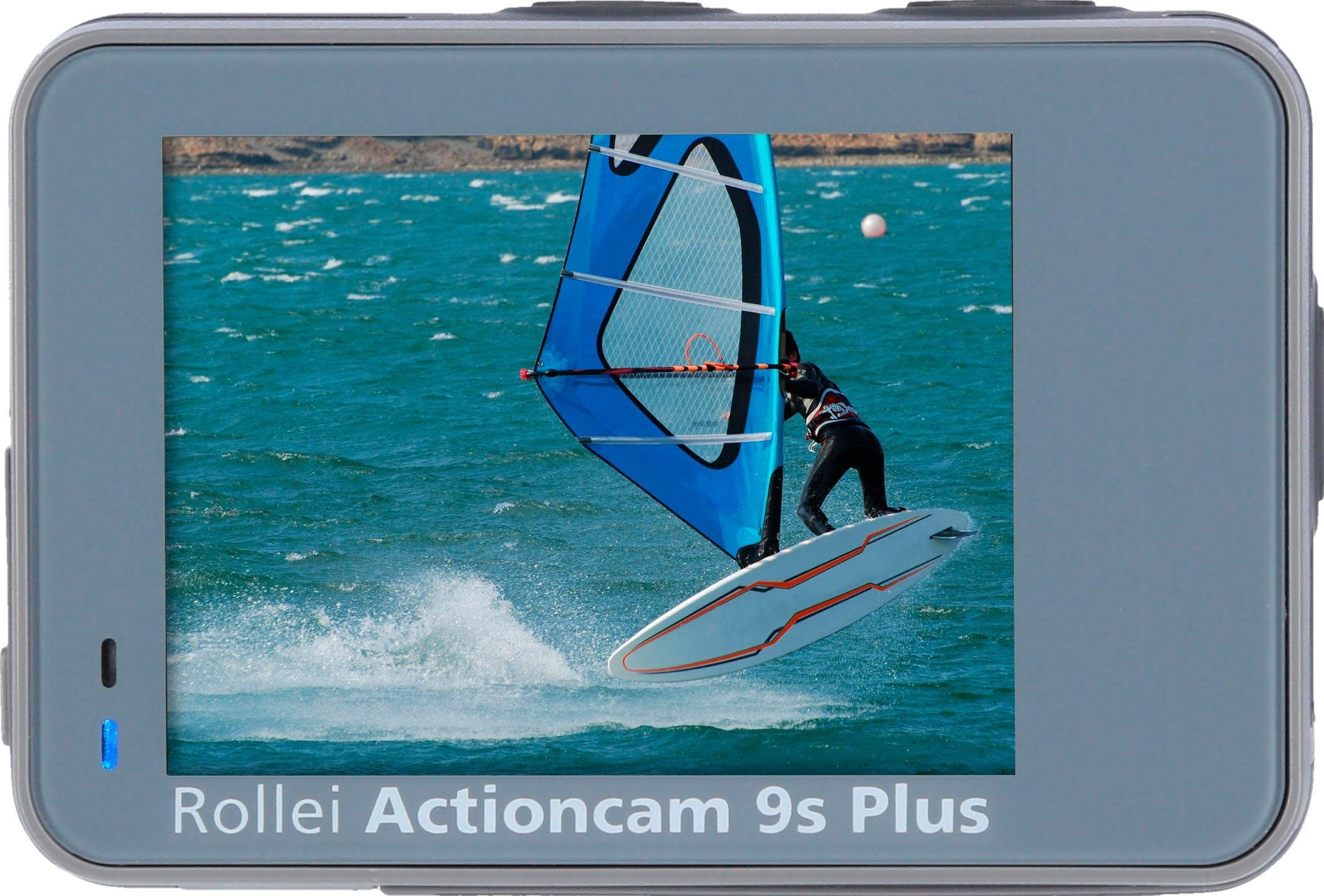 Actioncam mostra un windsurfista che salta sulle onde. Vela blu, tavola bianca e acqua blu. La fotocamera è grigia.