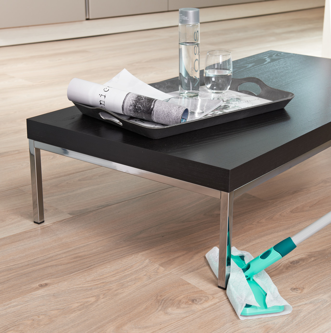 Ein schwarzer Couchtisch mit Tablett, Wasserflasche und Glas. Ein Mopp reinigt den Holzboden.